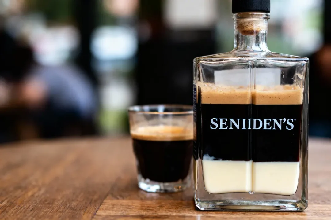 Bouteille transparente avec étiquette noire, liquide brun et crèmeux, posée sur une table en bois, arrière flou.