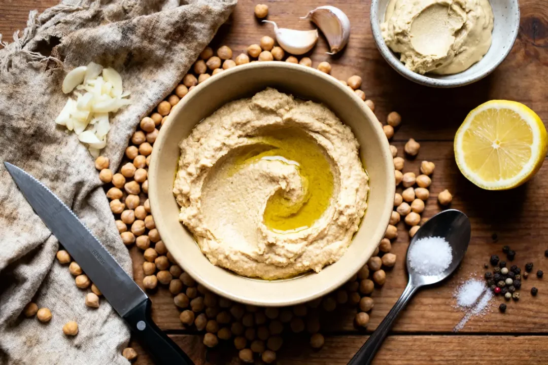 Bol de houmous avec huile d'olive, entouré de pois chiches, citron, ail, sel, poivre, couteau, tissu.