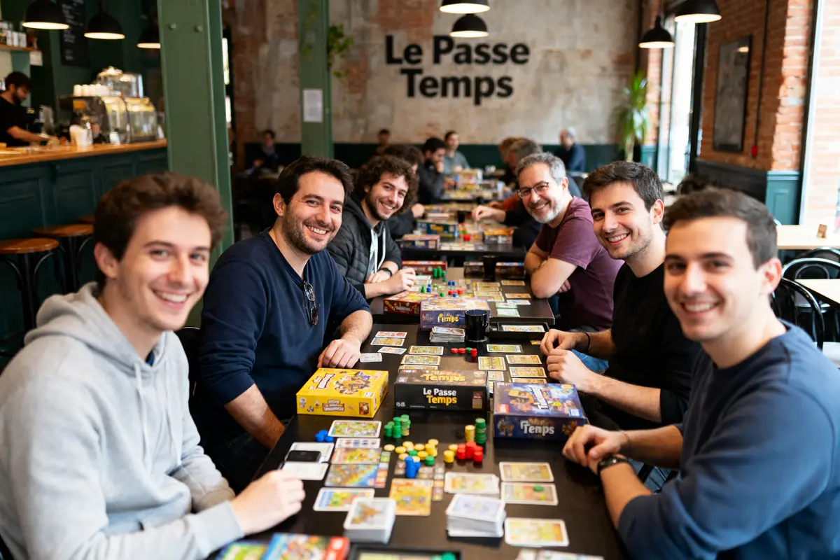 Un groupe d'hommes souriants joue à des jeux de société dans un café nommé "Le Passe Temps".