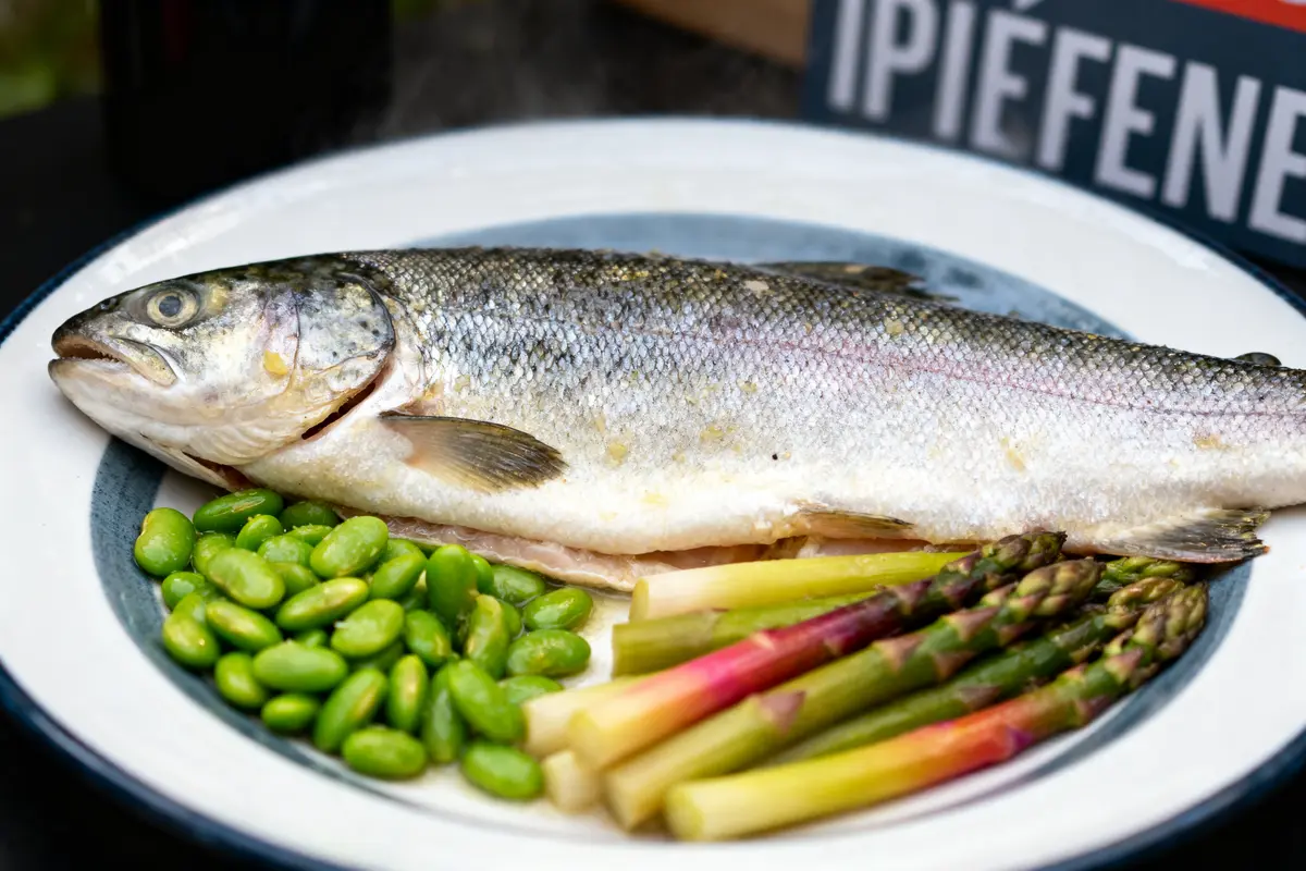 Poisson entier avec edamame et asperges sur assiette blanche, fond flou avec texte incomplètement visible.