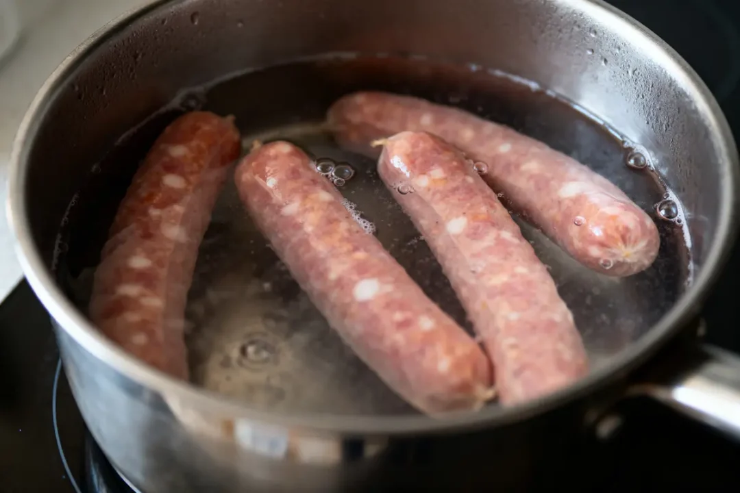 Quatre saucisses crues cuisent dans une casserole d'eau bouillante sur une cuisinière.