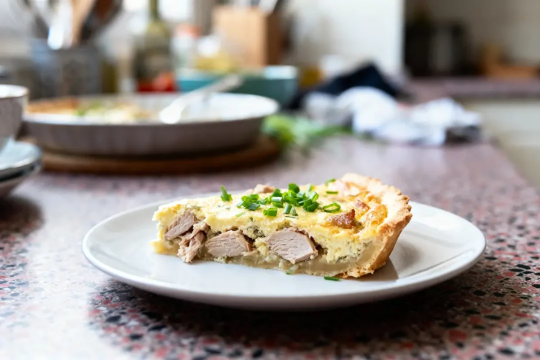 Part de quiche garnie de viande et ciboulette sur une assiette blanche, cuisine floue en arrière-plan.