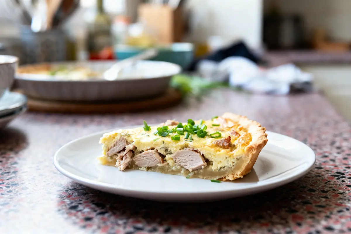 Part de quiche garnie de viande et ciboulette sur une assiette blanche, cuisine floue en arrière-plan.