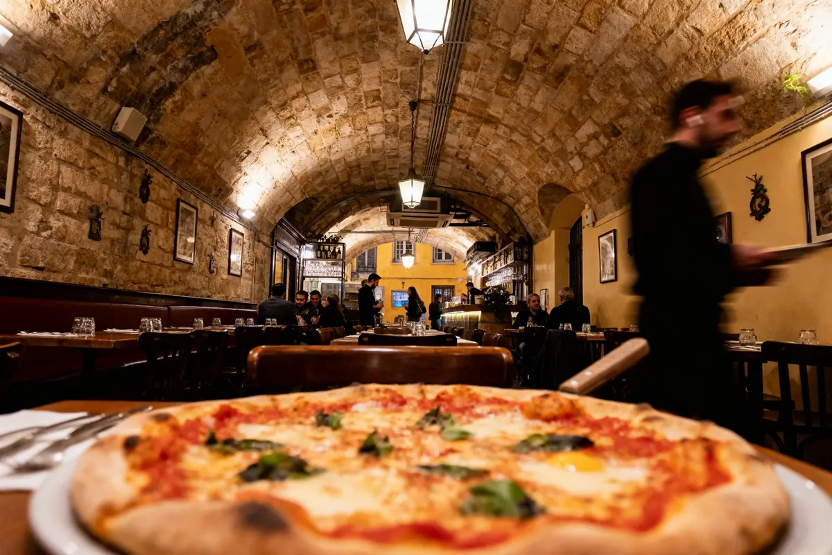 Pizza au premier plan dans un restaurant à voûtes avec des convives assis et serveurs debout.